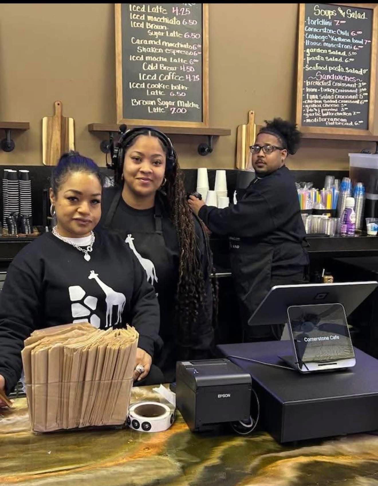 The Cornerstone Café team at the counter — warm brown interior with chalkboard menu